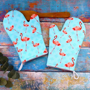 Könnte beinhalten: Ein Paar Ofenhandschuhe mit hellblauem Hintergrund und einem sich wiederholenden Muster aus rosa Flamingos. Die Handschuhe haben eine weiße Schlaufe zum Aufhängen und eine rosa Einfassung. Ein Holzlöffel liegt dahinter.