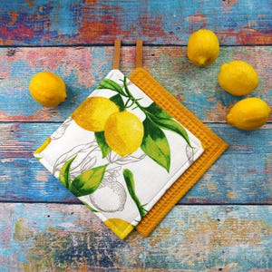 Könnte beinhalten: Zwei gelb-weiße Topflappen mit Zitronen- und Blattmuster und ein gelber Waffel-Topflappen. Die Topflappen haben braune Lederaufhängeschlaufen. Vier gelbe Zitronen sind um die Topflappen auf einer blau- und holzmaserierten Oberfläche verstreut.