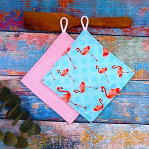 Könnte beinhalten: Zwei quadratische Topflappen. Einer ist rosa mit Waffelstruktur, der andere hellblau mit Flamingomuster. Die Flamingos sind rot und der Hintergrund hat kleine blaue Kreuze. Beide Topflappen haben weiße Aufhängeschlaufen.