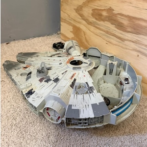 May include: A large, open, light gray and white toy spaceship with detailed panels, red and black accents, and a central circular cockpit. The toy has an open interior with seating and control panels. The toy is on a beige carpet.