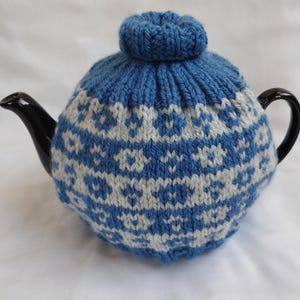 May include: A black teapot with a blue and white knitted tea cosy. The cosy features a repeating floral pattern in light blue and white. The cosy covers the teapot and has a small, round knitted top. The teapot has a black handle and spout.