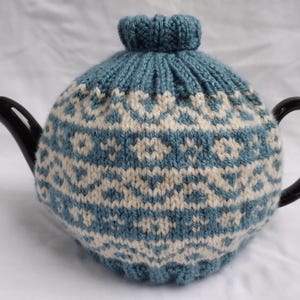 May include: A black teapot with a blue and cream knitted tea cosy. The cosy has a geometric pattern and covers the teapot. The teapot has a curved handle and spout. The cosy is a traditional design.