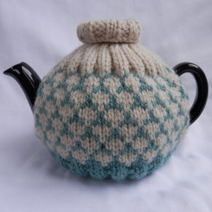 May include: A black teapot with a cream and teal knitted tea cosy. The cosy features a geometric pattern and a ribbed top. The teapot has a black handle and spout. The cosy is designed to keep the tea warm.