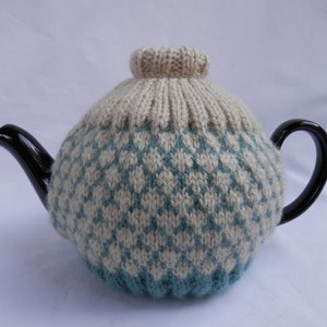 May include: A knitted teapot cosy in a cream and teal colour pattern. The cosy is made with a diamond stitch pattern and has a rounded top.