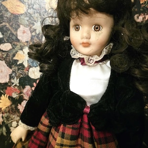 May include: A porcelain doll with dark brown hair, wearing a black velvet jacket, a white blouse with a lace collar, and a red and black plaid skirt.