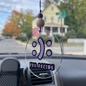 May include: A clear plastic car charm with a purple smiley face and the text "YOU DECIDE". The charm is hanging from a black string with purple and white beads.