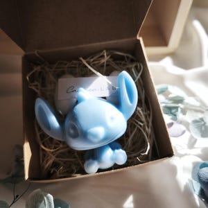 Può includere: Statuetta di Stitch azzurra all'interno di una scatola di cartone marrone piena di paglia. La statuetta ha grandi orecchie ed è in posizione seduta. È visibile una piccola etichetta bianca con testo. Fiori decorativi in primo piano.