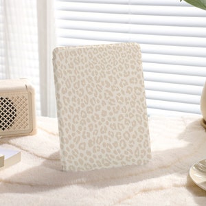 Könnte beinhalten: Ein beige-weißer Notizbuchumschlag mit Leopardenmuster. Der Umschlag ist aus einem weichen, strukturierten Material gefertigt und hat einen dezenten Glanz.