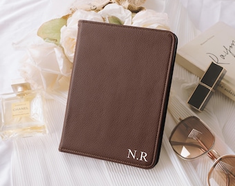 May include: A brown leather journal with the initials "N.R" in white. The journal is on a white surface with a perfume bottle, a book, sunglasses, and a lipstick. The journal is closed and has a textured surface.