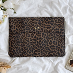 Pode incluir: Uma capa para laptop com estampa de leopardo marrom e preto com as iniciais douradas "KA" na frente. A capa é retangular e possui fecho de aba. O fundo inclui um arranjo floral branco, um livro e óculos de sol.