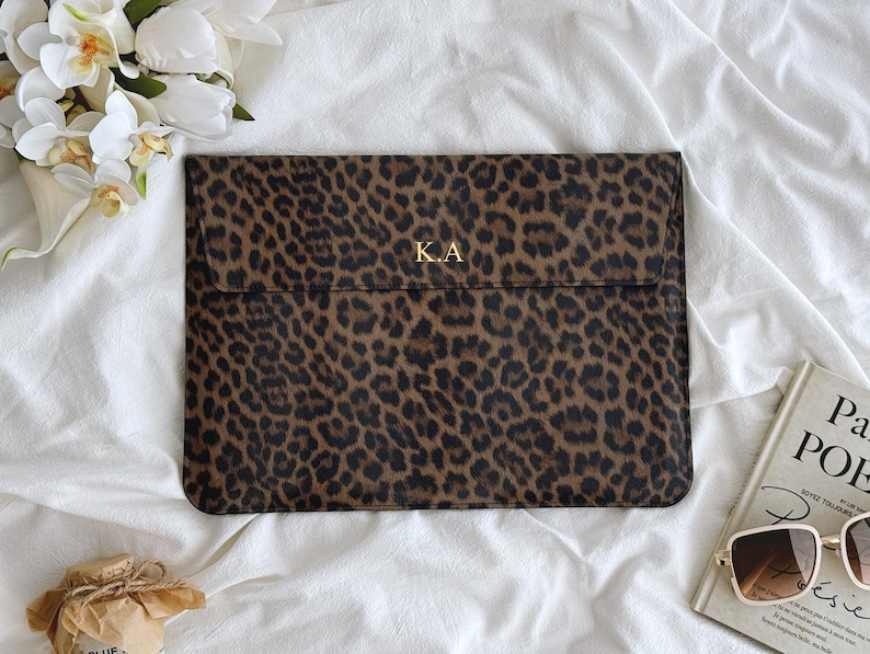 K&ouml;nnte beinhalten: Eine braun-schwarze Laptop-H&uuml;lle mit Leopardenmuster und den goldenen Initialen "KA" auf der Vorderseite. Die H&uuml;lle ist rechteckig und hat einen Klappverschluss. Im Hintergrund befinden sich ein wei&szlig;es Blumenarrangement, ein Buch und eine Sonnenbrille.