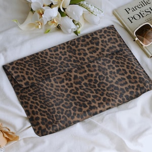 K&ouml;nnte beinhalten: Eine braun-schwarze Laptop-H&uuml;lle mit Leopardenmuster und Klappenverschluss. Die H&uuml;lle liegt auf einer wei&szlig;en Oberfl&auml;che neben einem Buch, einer Sonnenbrille und Blumen. Das Buch tr&auml;gt den Titel "Pareille PO&Eacute;SIE".