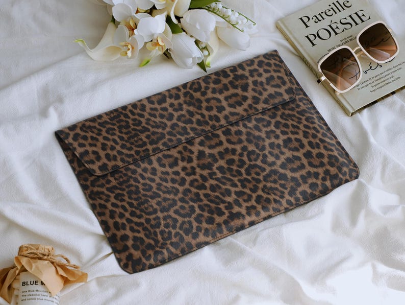 K&ouml;nnte beinhalten: Eine braun-schwarze Laptop-H&uuml;lle mit Leopardenmuster und Klappenverschluss. Die H&uuml;lle liegt auf einer wei&szlig;en Oberfl&auml;che neben einem Buch, einer Sonnenbrille und Blumen. Das Buch tr&auml;gt den Titel "Pareille PO&Eacute;SIE".