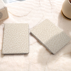 May include: Two beige e-reader covers with a leopard print design. The covers are on a white fuzzy surface.