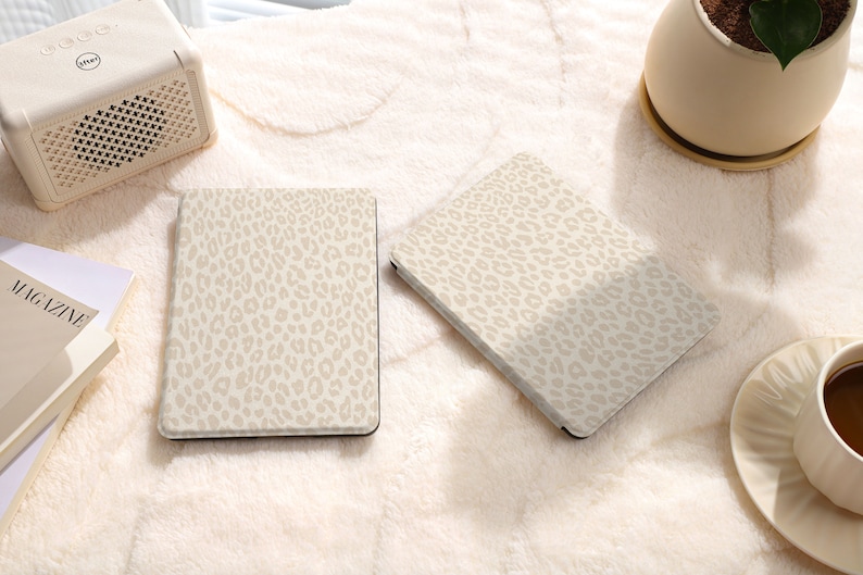 May include: Two beige e-reader covers with a leopard print design. The covers are on a white fuzzy surface.