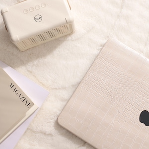 May include: A white laptop computer with a crocodile-embossed leather case. The laptop has a black Apple logo on the lid. The laptop is sitting on a white fluffy surface. A white speaker with a black "after" logo is in the upper left corner of the image. A magazine with the text "OH! MAGAZINE" is in the lower left corner of the image.