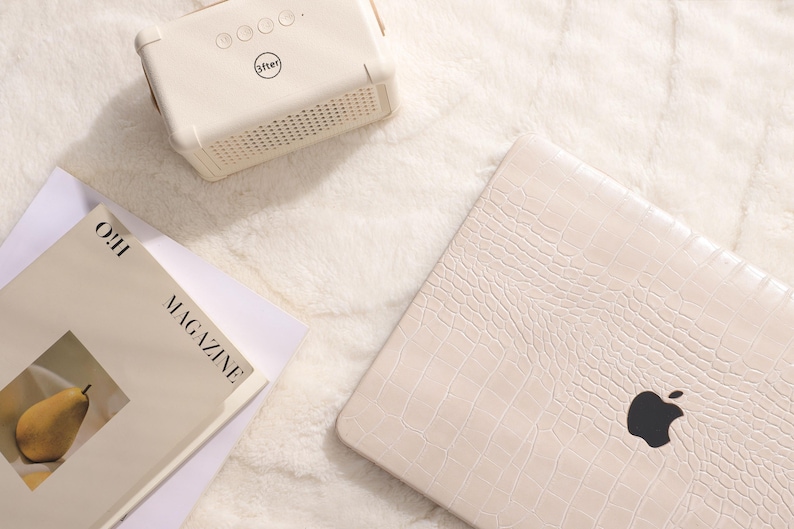 May include: A white laptop computer with a crocodile-embossed leather case. The laptop has a black Apple logo on the lid. The laptop is sitting on a white fluffy surface. A white speaker with a black "after" logo is in the upper left corner of the image. A magazine with the text "OH! MAGAZINE" is in the lower left corner of the image.