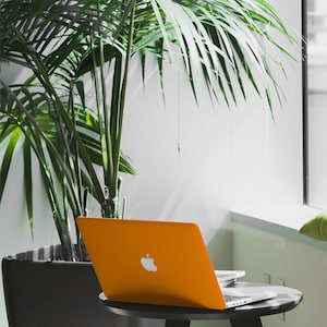 May include: A bright orange laptop computer with a white Apple logo on the lid. The laptop is sitting on a black table with a green plant in the background.