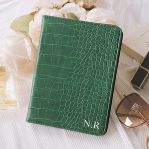 May include: A green, faux-crocodile textured journal with the initials "N.R" in white. The journal is rectangular and closed. Other items in the image include a perfume bottle, lipstick, sunglasses, and a book.