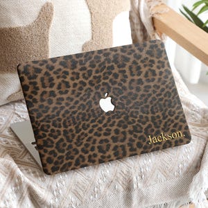 May include: A laptop with a leopard print cover featuring a white Apple logo. The cover is brown and black with the name "Jackson" in gold lettering. The laptop rests on a chair with a cream-colored cushion.