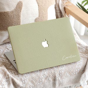 May include: A pale green laptop case with a ribbed texture. The Apple logo is cut out, and the name "Caroline" is written in cursive. The laptop is on a patterned blanket, with a cushion in the background.