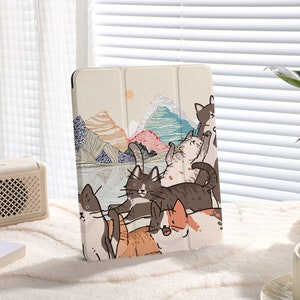 Puede incluir: Una funda para iPad de color crema con una ilustración colorida de gatos en un paisaje de montaña. Los gatos están en varias poses, algunos están sentados, otros de pie y otros saltando. El fondo es una paleta de colores suaves y pastel con una cordillera y un lago.
