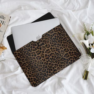 K&ouml;nnte beinhalten: Ein Laptop befindet sich in einer braunen und schwarzen Laptop-H&uuml;lle mit Leopardenmuster. Die H&uuml;lle ist ge&ouml;ffnet und zeigt den silbernen Laptop. Ein Buch, eine Sonnenbrille und ein kleines Glas befinden sich neben dem Laptop. Rechts ist ein Strau&szlig; wei&szlig;er Blumen.