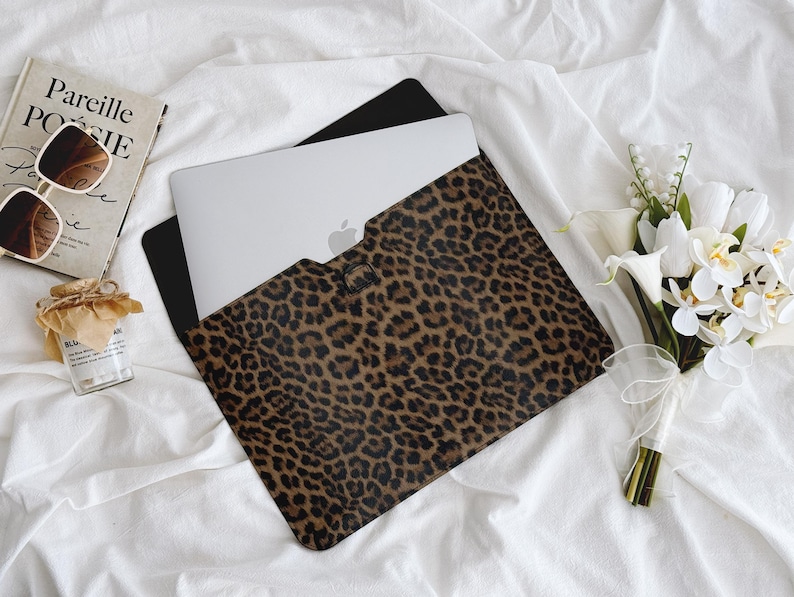 K&ouml;nnte beinhalten: Ein Laptop befindet sich in einer braunen und schwarzen Laptop-H&uuml;lle mit Leopardenmuster. Die H&uuml;lle ist ge&ouml;ffnet und zeigt den silbernen Laptop. Ein Buch, eine Sonnenbrille und ein kleines Glas befinden sich neben dem Laptop. Rechts ist ein Strau&szlig; wei&szlig;er Blumen.