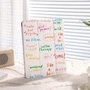 Pode incluir: Capa de tablet branca com frases manuscritas coloridas como "cute but psycho", "girl power" e "good vibes". A capa está sobre uma superfície branca perto de um alto-falante, livros e uma xícara de café.