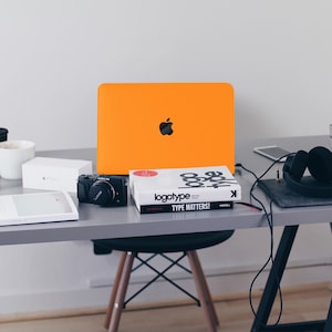 May include: A workspace desk with a laptop, camera, book, headphones, and a white box. The laptop is orange and has an Apple logo. The book is titled "logotype TYPE MATTERS!" The headphones are black and are connected to a phone.