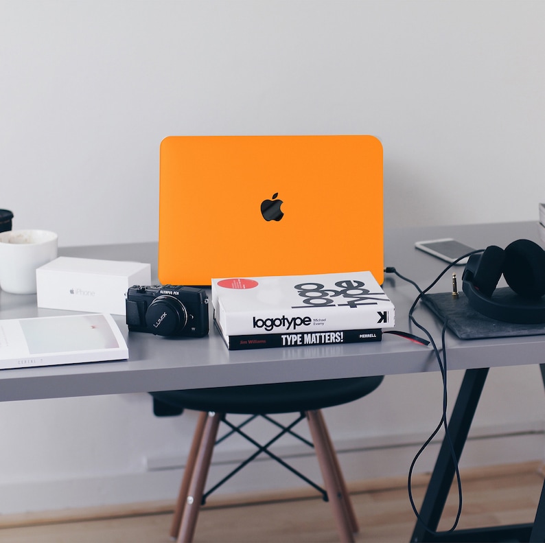 May include: A workspace desk with a laptop, camera, book, headphones, and a white box. The laptop is orange and has an Apple logo. The book is titled "logotype TYPE MATTERS!" The headphones are black and are connected to a phone.