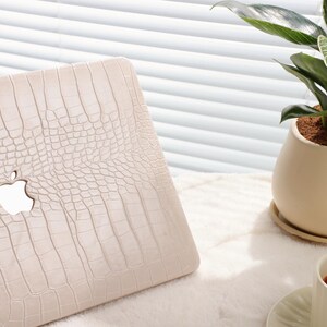 May include: A white laptop case with a crocodile skin pattern and the Apple logo. The case is on a white surface with a potted plant and a cup of tea in the background.