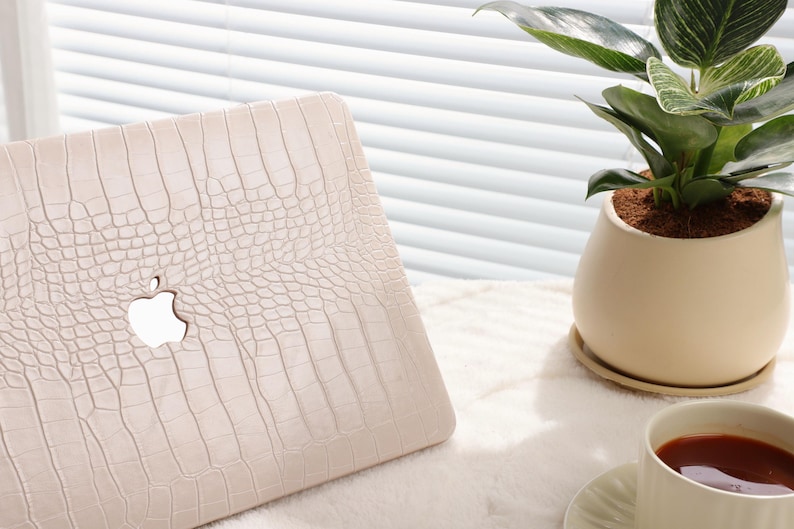 May include: A white laptop case with a crocodile skin pattern and the Apple logo. The case is on a white surface with a potted plant and a cup of tea in the background.