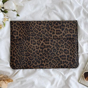 K&ouml;nnte beinhalten: Eine rechteckige Laptop-H&uuml;lle mit einem Leopardenmuster in Braun- und Schwarzt&ouml;nen. Die H&uuml;lle wird mit einer Klappe verschlossen. Der Hintergrund umfasst wei&szlig;en Stoff, ein Buch, eine Sonnenbrille und Blumen.