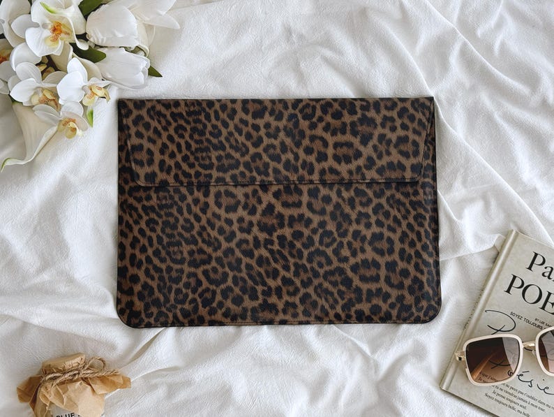 K&ouml;nnte beinhalten: Eine rechteckige Laptop-H&uuml;lle mit einem Leopardenmuster in Braun- und Schwarzt&ouml;nen. Die H&uuml;lle wird mit einer Klappe verschlossen. Der Hintergrund umfasst wei&szlig;en Stoff, ein Buch, eine Sonnenbrille und Blumen.