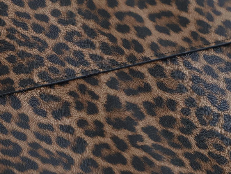 K&ouml;nnte beinhalten: Nahaufnahme eines braunen und schwarzen Leopardenmuster. Das Muster weist dunkelbraune Flecken auf hellem braunen Hintergrund auf. Eine schwarze Naht verl&auml;uft &uuml;ber das Bild und teilt das Muster.