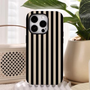 Pode incluir: Uma capa de telefone listrada em preto e bege. A capa tem um padrão de listras verticais e um recorte para a câmera. A capa do telefone é mostrada em frente a um alto-falante e uma planta em vaso.