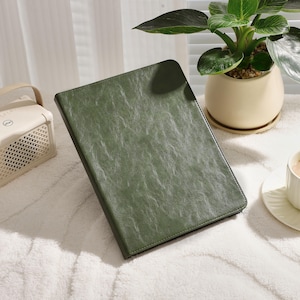 May include: A dark green leather-bound notebook or journal with a textured surface. The notebook is lying on a white fluffy surface.
