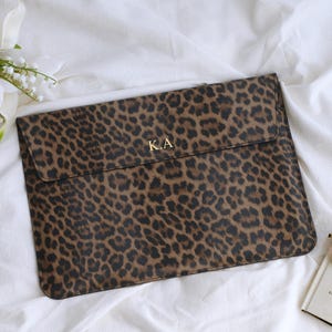 K&ouml;nnte beinhalten: Eine braun-schwarze Laptop-H&uuml;lle mit Leopardenmuster und den goldenen Initialen "K.A." auf der Vorderseite. Die H&uuml;lle ist rechteckig mit einem Klappenverschluss. Der Hintergrund umfasst wei&szlig;e Blumen, eine Sonnenbrille und ein Buch.