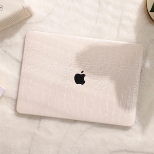 May include: A white laptop computer with a crocodile-embossed design and a black Apple logo. The laptop is on a white fuzzy surface.