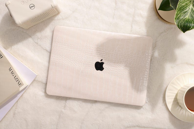 May include: A white laptop computer with a crocodile-embossed design and a black Apple logo. The laptop is on a white fuzzy surface.
