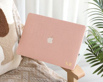 May include: A light pink laptop case with a crocodile skin texture. The case has a silver Apple logo and gold initials "S.R." The laptop case is resting on a wooden chair.