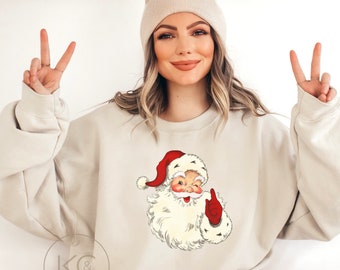 May include: Sand-colored sweatshirt featuring a vintage Santa Claus graphic with a red hat and gloves. The sweatshirt has the text "For oversized look order 1-2 sizes up" at the top. Below the Santa graphic are two red footprints. The sweatshirt is a neutral color.