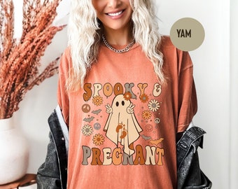 May include: Orange t-shirt with the text "Spooky & Pregnant" in brown and orange lettering. The shirt features a ghost graphic with peace signs, flowers, and bats. The shirt is a comfortable, oversized fit.