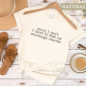May include: A white t-shirt with the text "sorry I can't have to feed my sourdough starter" printed on it. The shirt is on a wooden surface with a straw hat, sandals, and a cup of coffee.