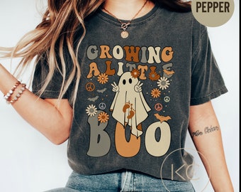 May include: A dark gray t-shirt with a vintage-style graphic of a ghost with the text "Growing a Little Boo". The graphic features peace signs, bats, and flowers in shades of orange, brown, and yellow.