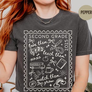 May include: Charcoal grey t-shirt with a white graphic. The design, within a stamp-like border, says "SECOND GRADE" and "2025", with school-themed illustrations and the phrases "love them", "teach them", and "watch them grow".