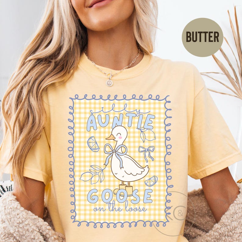 May include: Butter-colored t-shirt with a whimsical design featuring a cartoon goose wearing a bow. The text reads "Silly Auntie Goose on the Loose" in blue lettering, set against a yellow and white checkered background. The shirt has a crew neck.