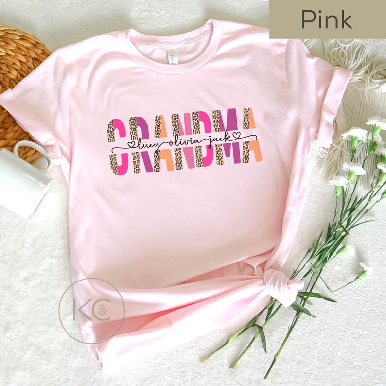 May include: Pink t-shirt with the word "Grandma" in colorful letters with a leopard print design. The names "Lucy, Olivia, Jack" are written in a smaller font below the word "Grandma".