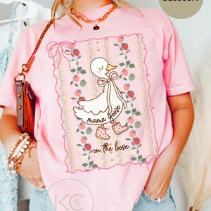 May include: Pink t-shirt with a graphic of a cartoon goose wearing boots and a bow. The text "mama goose on the loose" is printed below the goose. The shirt has a floral and strawberry design around the graphic.